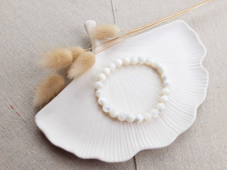 K&ouml;nnte beinhalten: Ein Armband aus wei&szlig;en Muschelkernperlen auf einer wei&szlig;en Keramikplatte mit getrockneten Blumen.