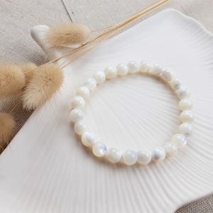 K&ouml;nnte beinhalten: Ein Armband aus wei&szlig;en Muschelkernperlen auf einem wei&szlig;en Teller mit getrockneten Blumen.