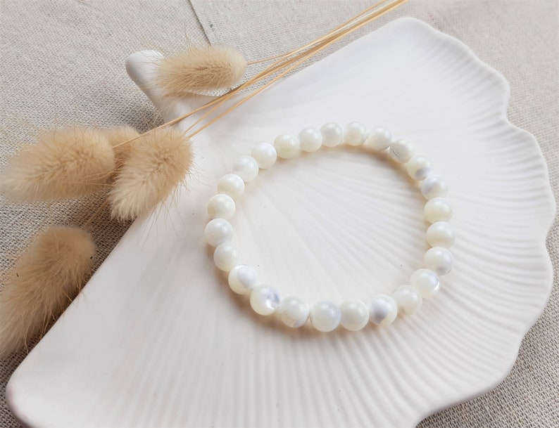 K&ouml;nnte beinhalten: Ein Armband aus wei&szlig;en Muschelkernperlen auf einem wei&szlig;en Teller mit getrockneten Blumen.