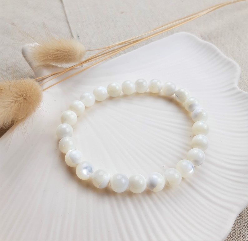 K&ouml;nnte beinhalten: Ein wei&szlig;es Muschelperlenarmband mit glatter, irisierender Oberfl&auml;che. Das Armband besteht aus nat&uuml;rlichen Muschelperlen und ist auf einer durchsichtigen Gummikordel aufgef&auml;delt.