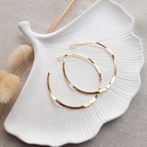 May include: A pair of gold hoop earrings with a hammered texture. The earrings are displayed on a white ceramic dish.