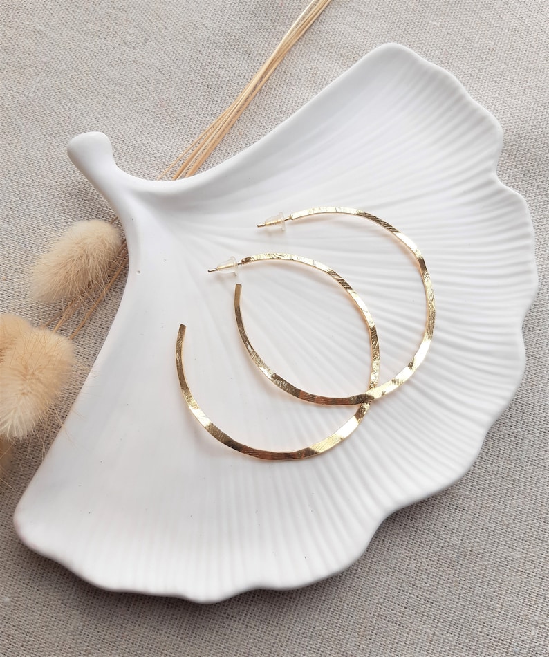 May include: A pair of gold hoop earrings with a hammered texture. The earrings are displayed on a white ceramic dish.