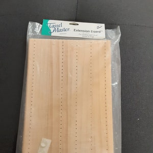 May include: A wooden extension board for creating super-sized tassels and fringe. The board is light brown and has rows of holes for threading yarn. It comes with adhesive pads for attaching to a surface.