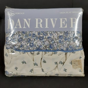 May include: A white twin flat sheet with a blue floral pattern and a ruffled edge. The sheet is packaged and labeled "Dan River" with the words "Twin Flat Sheet" and "100% Cotton Percale".