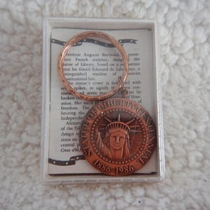 May include: A copper-colored Statue of Liberty Centennial medallion and a copper ring are displayed in a clear plastic case. The medallion features the Statue of Liberty and the dates 1886-1986. Text about the statue is visible in the background.