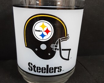 Puede incluir: Un vaso de vidrio transparente con un fondo blanco esmerilado. El vaso presenta un logotipo de casco de fútbol americano de los Steelers de Pittsburgh en negro y amarillo y la palabra "Steelers" en letras negras. El vaso es adecuado para bebidas.