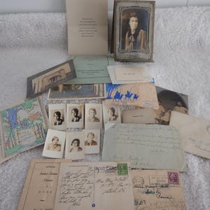 May include: A collection of vintage ephemera, including postcards, photographs, and letters, arranged on a white surface. The items vary in size and shape, with some featuring handwritten text and illustrations. A framed portrait and a small booklet are also present.