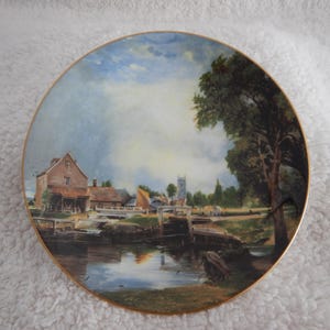 May include: A decorative plate featuring a scenic landscape painting. The artwork depicts a river scene with buildings, a church steeple, and trees under a cloudy sky. The plate has a gold-colored rim.