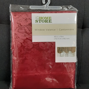 May include: A red window valance in a clear plastic package. The valance is 60 inches long and 19 inches wide, with a floral pattern. The packaging includes the text "the HOME STORE" and "Window Valance | Cantonnière".