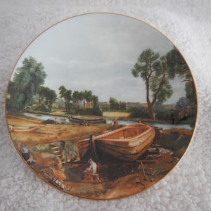May include: Decorative plate featuring a detailed landscape painting of a river scene with boats and figures. The plate has a gold rim and a classic aesthetic, suitable for display or collection.