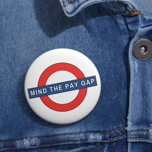 May include: A white button with a red circle and blue text that reads "MIND THE PAY GAP". The button is a parody of the London Underground sign.
