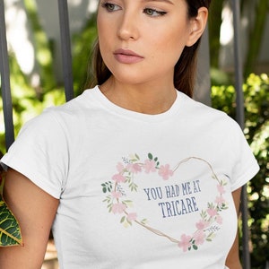 Op de afbeelding: Wit T-shirt met een bloemenhartontwerp en de tekst "YOU HAD ME AT TRICARE". Het shirt wordt gecombineerd met roze en wit gestreepte shorts met een strik in de taille. Het model leunt tegen een metalen hek.