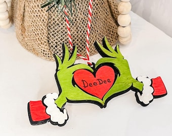 Puede incluir: Un adorno navideño de madera con manos verdes que forman un corazón alrededor de un corazón rojo con el nombre "DeeDee" en blanco. Las manos tienen puños rojos y ribete de piel blanca. El adorno cuelga de una cuerda roja y blanca.