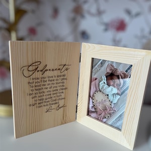 May include: A wooden photo frame with a light-colored finish, open to display a baby photo. The left side of the frame has engraved text that reads "Godparents." The right side holds a photo of a baby with a wooden name tag.