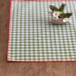 Könnte beinhalten: Ein grün-weiß kariertes Platzset mit rosa Rand liegt auf einer Holzoberfläche. Ein kleiner, rosa Vogel-Keramiktopf, gefüllt mit Grün und kleinen weißen Blumen, steht auf dem Platzset. Die Szene wirkt rustikal und frühlingshaft.
