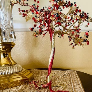 May include: A wire-wrapped tree sculpture with red and gold wire, adorned with red and blue beads and gold leaf accents. The tree is displayed on a patterned table runner.