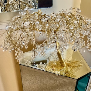 May include: A gold wire sculpture of a weeping willow tree with clear crystal accents. The tree is displayed on a mirrored pedestal.