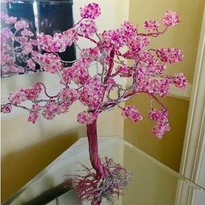 Op de afbeelding: Een roze kralen draadboom sculptuur met een zilveren draad basis. De boom is gemaakt van draad en kralen en heeft een natuurlijke, organische uitstraling.