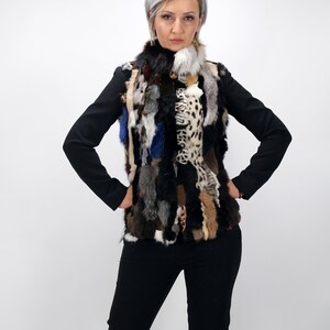 Può includere: Una donna indossa una giacca nera con un gilet di pelliccia multicolore. Il gilet è fatto di diversi tipi di pelliccia, tra cui leopardo, volpe e visone. La donna è in piedi con le mani sui fianchi.