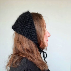 May include: A black knitted headscarf with a triangular shape and a textured ribbed pattern. The headscarf features thin black ties that hang down from the sides, allowing it to be secured under the chin with a simple knot.