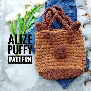 Könnte beinhalten: Eine braune gehäkelte Tragetasche mit einem Hirschgesichtsdesign. Die Tasche hat zwei braune Ohren, eine braune Nase und ein braunes und weißes Muster. Die Tasche liegt auf einer blauen Jeans und einer weißen Betonfläche. Der Text "Alize Puffy Pattern" ist im Bild sichtbar.