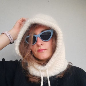 May include: A white knitted hooded scarf with a long tie. The scarf is made of a soft, fluffy yarn and has a relaxed, casual look. The model is wearing the scarf with a black shirt and blue cat-eye sunglasses.