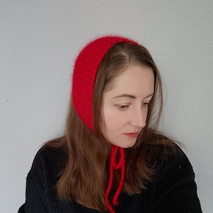Könnte beinhalten: Eine rote Strickmütze mit einer langen Schleife. Die Mütze ist aus weichem, flauschigem Material und hat ein schlichtes Design.