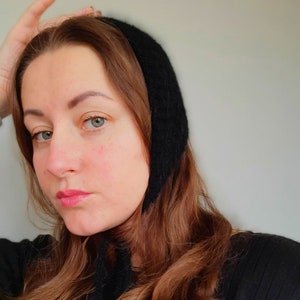 May include: A woman with long brown hair is wearing a black knit hat with a long tie. The hat covers her ears and the tie hangs down her neck. She is wearing a black shirt.