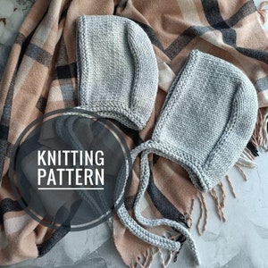 May include: Two grey knitted baby bonnets with ties, lying on a brown and black checked blanket. The text "Knitting Pattern" is visible in a circle.