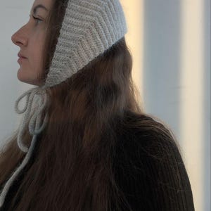 May include: A light gray, knitted bonnet featuring a textured chevron pattern and a ribbed edge. The headwear is secured under the chin with two matching knitted ties that hang down, providing a snug fit around the head and ears.