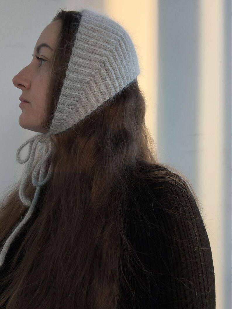 May include: A light gray, knitted bonnet featuring a textured chevron pattern and a ribbed edge. The headwear is secured under the chin with two matching knitted ties that hang down, providing a snug fit around the head and ears.