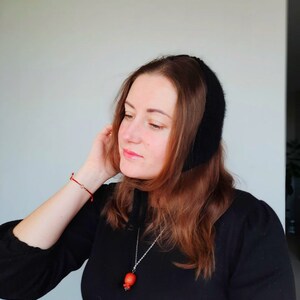 May include: A black knit bonnet with a soft texture, paired with a silver-toned necklace featuring a small, round, orange-red pendant. The bonnet covers the top of the head and ties under the chin, while the necklace rests against a black top.