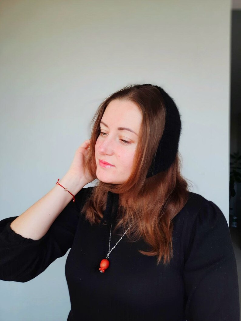 May include: A black knit bonnet with a soft texture, paired with a silver-toned necklace featuring a small, round, orange-red pendant. The bonnet covers the top of the head and ties under the chin, while the necklace rests against a black top.