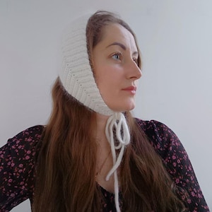May include: A white knitted bonnet featuring a ribbed texture and long yarn ties that fasten under the chin. The headwear is crafted from a soft, fuzzy yarn and covers the top and sides of the head, providing a snug fit around the face and ears.