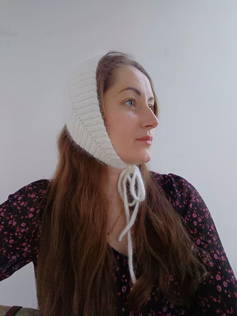 May include: A white knitted bonnet featuring a ribbed texture and long yarn ties that fasten under the chin. The headwear is crafted from a soft, fuzzy yarn and covers the top and sides of the head, providing a snug fit around the face and ears.