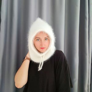 May include: A white, fluffy balaclava-style hat with a drawstring closure. The hat is made of a soft, fuzzy material and covers the head and neck. The person in the image is wearing a black shirt.