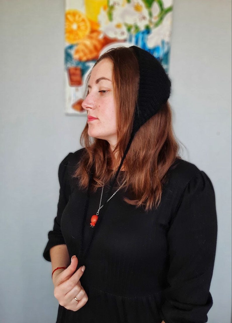May include: A black knitted bonnet with long ties that drape down the front. The bonnet features a textured, ribbed stitch pattern and covers the top and back of the head, secured with thin black yarn ties that hang loosely over the chest.
