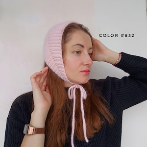 May include: A light pink knitted bonnet with a ribbed texture and long chin ties. The bonnet features a soft, fuzzy appearance and is labeled with the text COLOR #832. The design covers the top and sides of the head, securing under the chin.
