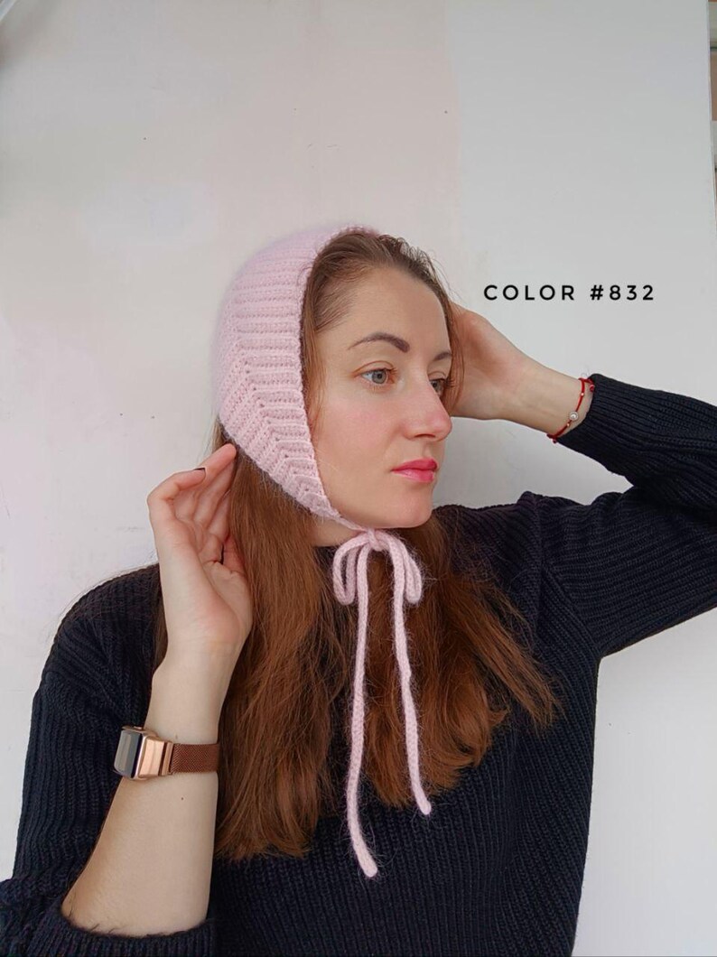 May include: A light pink knitted bonnet with a ribbed texture and long chin ties. The bonnet features a soft, fuzzy appearance and is labeled with the text COLOR #832. The design covers the top and sides of the head, securing under the chin.