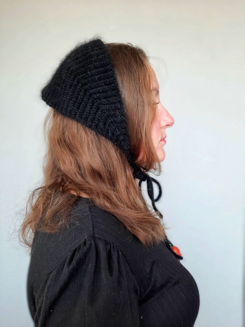 May include: A black knitted headscarf with a triangular shape and a textured ribbed pattern. The headscarf features thin black ties that hang down from the sides, allowing it to be secured under the chin with a simple knot.