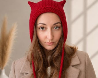 May include: A bright red knitted beanie featuring two pointed cat-like ears on the top and two long, matching red knitted ties hanging down from the sides. The hat is constructed with a textured knit pattern and covers the crown of the head.