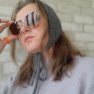 May include: A dark gray knitted bonnet with tie strings under the chin, paired with gold-rimmed sunglasses featuring brown lenses and an orange beaded ring. The bonnet has a textured, fuzzy appearance and is worn over light brown hair.