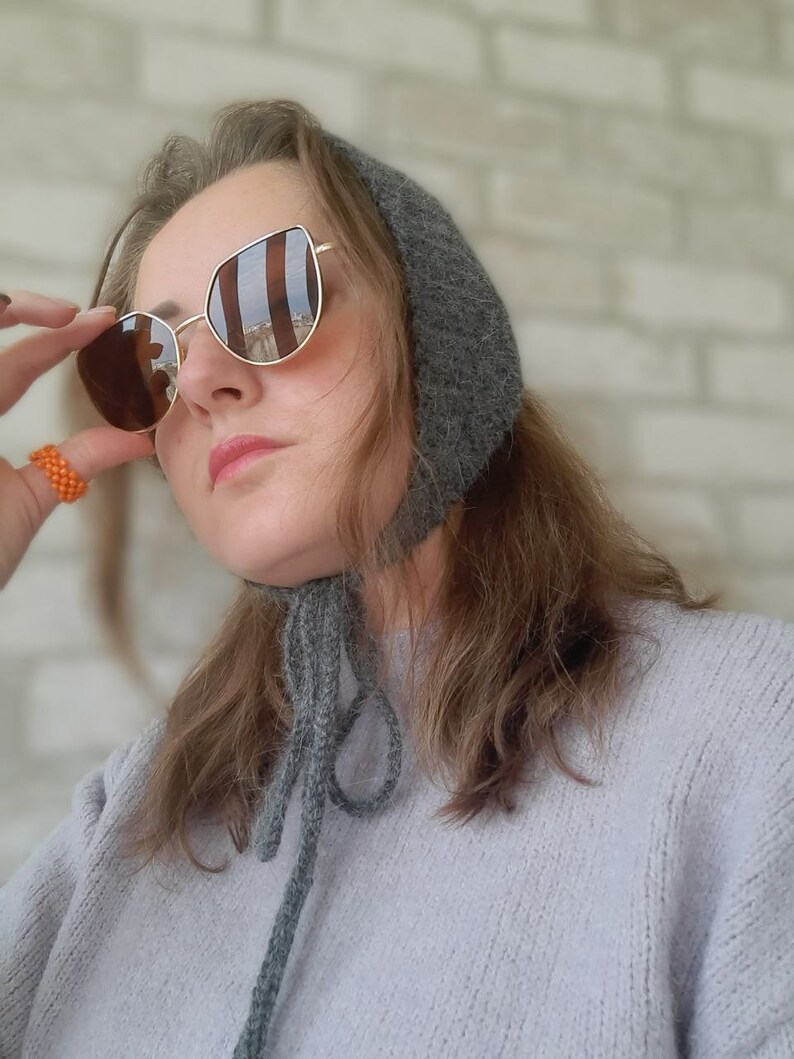 May include: A dark gray knitted bonnet with tie strings under the chin, paired with gold-rimmed sunglasses featuring brown lenses and an orange beaded ring. The bonnet has a textured, fuzzy appearance and is worn over light brown hair.