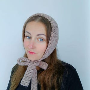May include: A woman wearing a light brown knitted bonnet with a long tie that is tied in a bow at the neck.