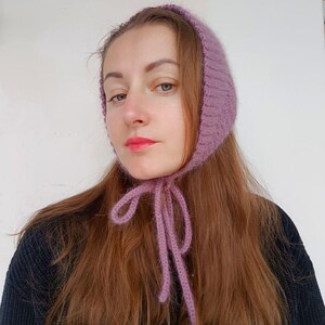 May include: A mauve-colored knitted bonnet featuring a textured, ribbed pattern and long, thin knitted ties that fasten under the chin. The headwear is crafted from a soft, fuzzy yarn and frames the face with a snug, rounded fit.