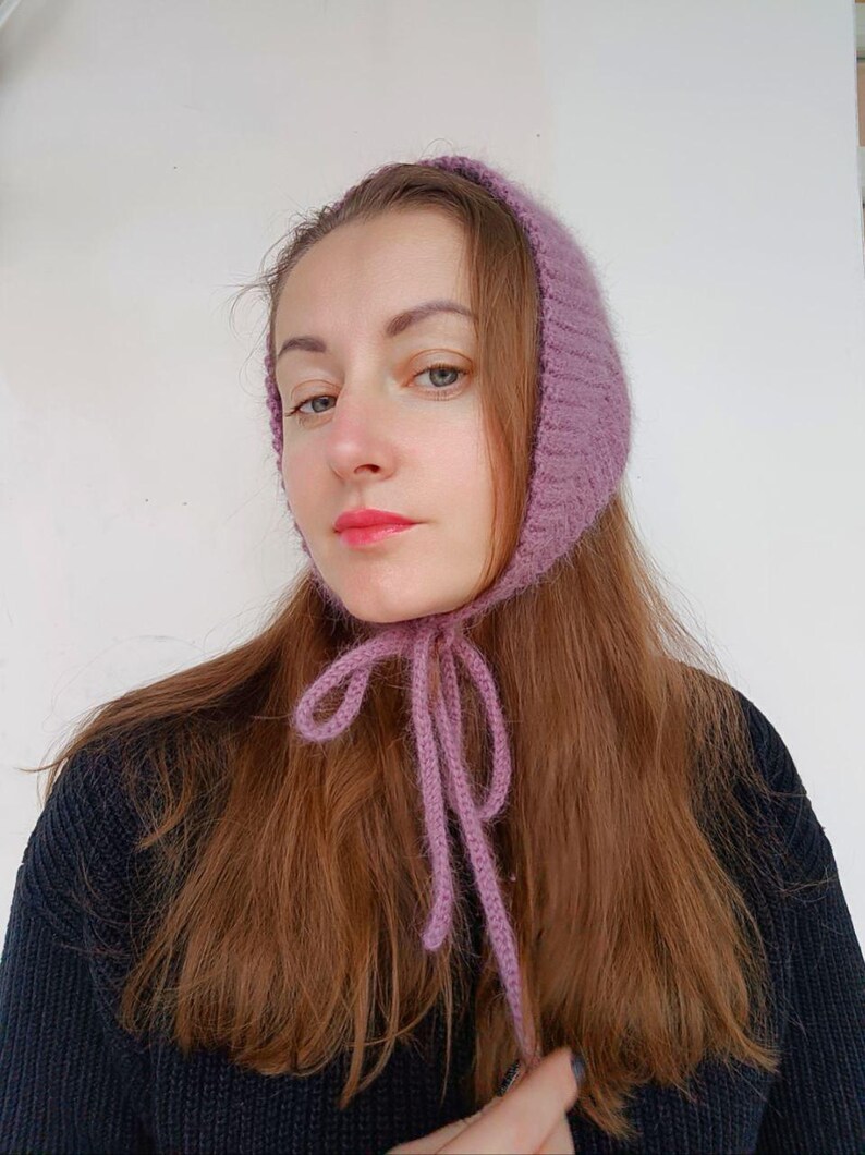 May include: A mauve-colored knitted bonnet featuring a textured, ribbed pattern and long, thin knitted ties that fasten under the chin. The headwear is crafted from a soft, fuzzy yarn and frames the face with a snug, rounded fit.