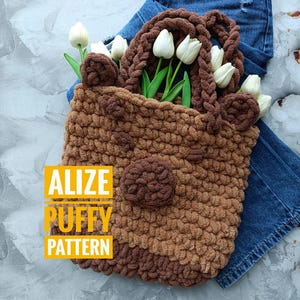 Puede incluir: Una bolsa de mano de crochet marrón y beige con orejas de oso y una nariz de pompón marrón. La bolsa está llena de tulipanes blancos y el texto "Alize Puffy Pattern" es visible en la esquina inferior izquierda.