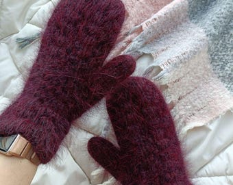 May include: A pair of burgundy mittens made from a fuzzy, textured material. The mittens are on a white quilted surface with a patterned scarf in the background. These mittens are a warm winter accessory.