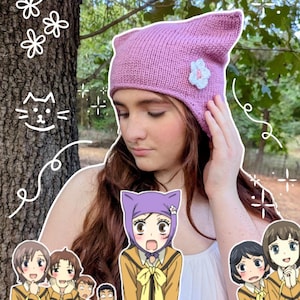 May include: A light purple knitted cat-ear beanie featuring a small white flower appliqué on the side. The hat has a pointed top and a textured knit pattern, worn as a head accessory with a soft, flexible fabric construction.