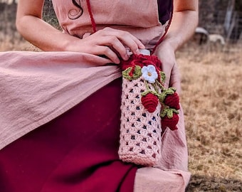 May include: A woman wearing a burgundy dress and a light pink apron dress, with a matching crochet headband. She is holding a crochet water bottle holder with red roses and strawberries. The background is a field with trees.